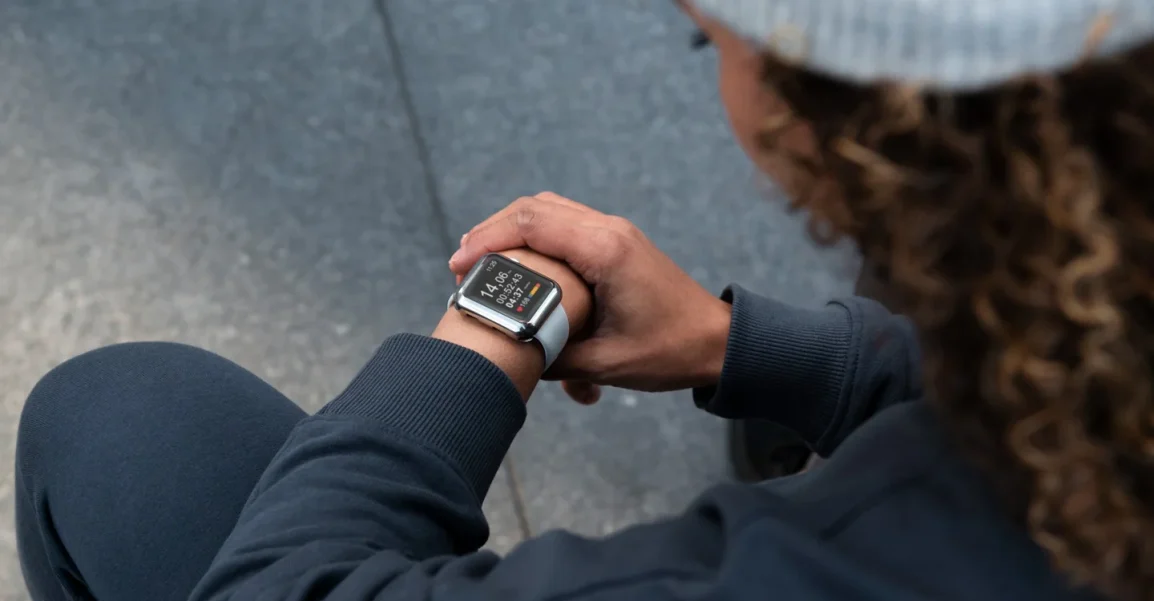 Person seated, wearing a beanie and dark hoodie, looking at a smartwatch on their left wrist.