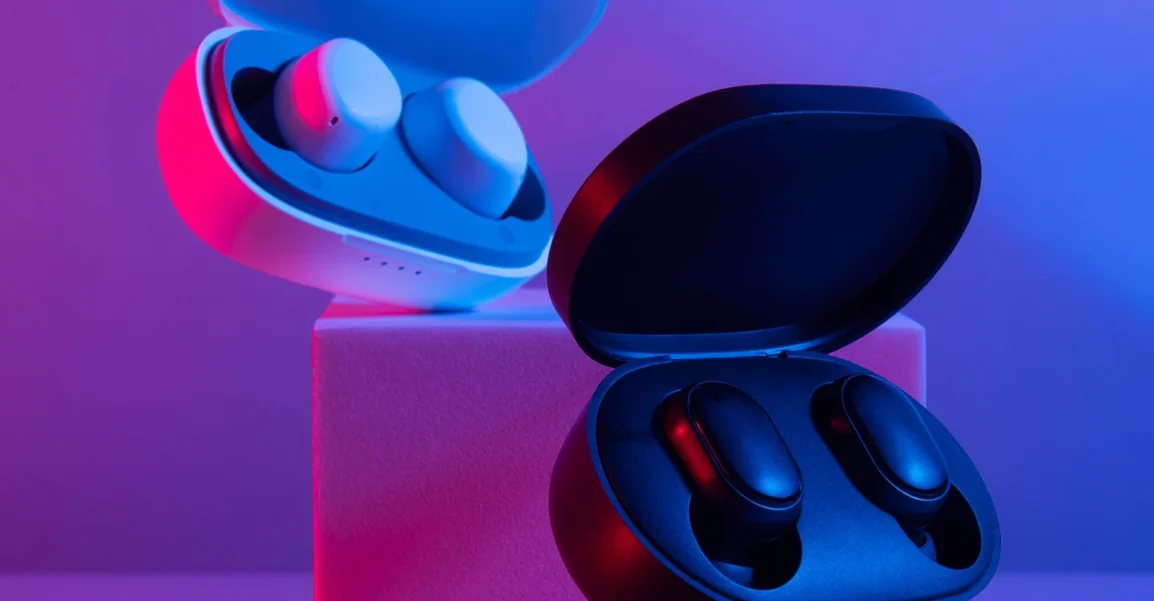 Open white wireless earbud case with white earbuds resting on a pink pedestal; a black earbud case with black earbuds sits on a blue-pink platform.