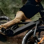 Cyclist in tan pants crashes on a black mountain bike on a rocky dirt trail.