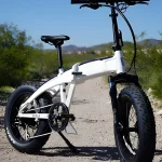 White electric bike on a dirt trail with desert shrubs and distant mountains under a clear blue sky.
