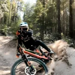 Mountain biker in a blue helmet leans into a dirt turn on a forest trail, kicking up dusted debris behind him/her.